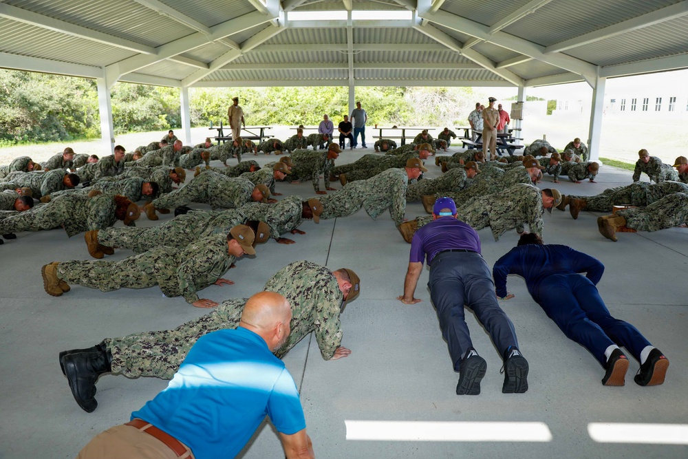 A large group of military personnel in camouflage uniforms are performing push-ups under an open-air pavilion structure. Several civilians in casual clothing are also participating alongside the uniformed personnel, suggesting a military orientation or community engagement event. A few individuals in uniform are standing and observing the exercise.