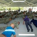 A large group of military personnel in camouflage uniforms are performing push-ups under an open-air pavilion structure. Several civilians in casual clothing are also participating alongside the uniformed personnel, suggesting a military orientation or community engagement event. A few individuals in uniform are standing and observing the exercise.