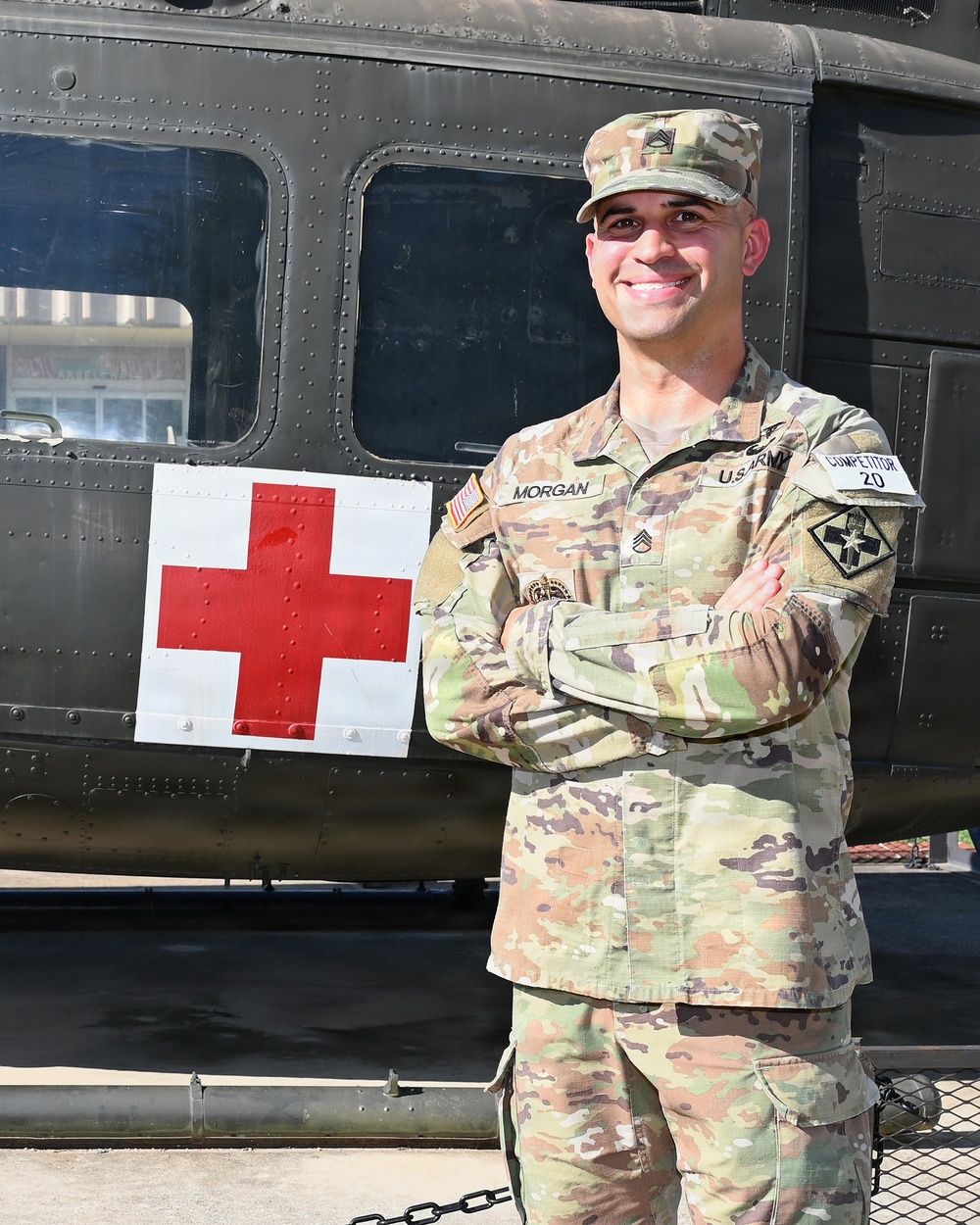 A U.S. Army soldier identified by name tape as 'Morgan' stands with arms crossed in front of a military medical helicopter marked with a Red Cross symbol. The soldier wears OCP (Operational Camouflage Pattern) uniform with Staff Sergeant rank insignia on the cap and a '2nd Infantry Division' patch on the left shoulder. A 'COMPETITOR 20' tab is visible on the right sleeve, suggesting participation in a military competition.