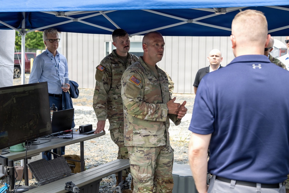 A military briefing or demonstration is taking place outdoors under a blue canopy tent. Two U.S. Army soldiers in OCP (Operational Camouflage Pattern) uniforms appear to be presenting or explaining something to a mixed group of military and civilian personnel. A laptop and monitor are set up on a folding table, suggesting a technology or equipment demonstration.