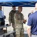 A military briefing or demonstration is taking place outdoors under a blue canopy tent. Two U.S. Army soldiers in OCP (Operational Camouflage Pattern) uniforms appear to be presenting or explaining something to a mixed group of military and civilian personnel. A laptop and monitor are set up on a folding table, suggesting a technology or equipment demonstration.