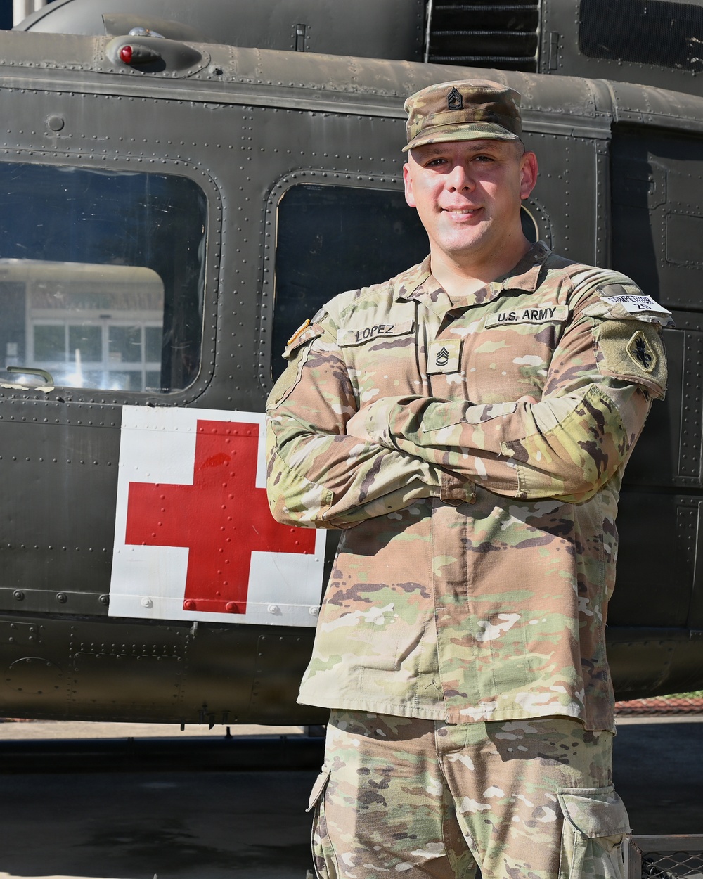 A U.S. Army Staff Sergeant named Lopez stands with arms crossed in front of a military medical helicopter marked with a Red Cross symbol. He is wearing OCP (Operational Camouflage Pattern) uniform with visible rank insignia and name tape. The image appears to be an official or semi-official portrait-style photograph taken outdoors.