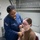 A U.S. Navy medical personnel is conducting a medical examination on a seated patient using a stethoscope. The examiner is wearing a navy blue U.S. Navy uniform with an American flag patch and a rating badge, while the patient is seated on a medical examination chair wearing a tan/brown t-shirt. The setting appears to be a military medical bay or sick bay facility.