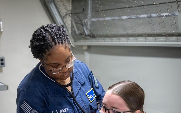 A U.S. Navy medical personnel is conducting a medical examination on a seated patient using a stethoscope. The examiner is wearing a navy blue U.S. Navy uniform with an American flag patch and a rating badge, while the patient is seated on a medical examination chair wearing a tan/brown t-shirt. The setting appears to be a military medical bay or sick bay facility.
