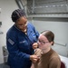 A U.S. Navy medical personnel is conducting a medical examination on a seated patient using a stethoscope. The examiner is wearing a navy blue U.S. Navy uniform with an American flag patch and a rating badge, while the patient is seated on a medical examination chair wearing a tan/brown t-shirt. The setting appears to be a military medical bay or sick bay facility.