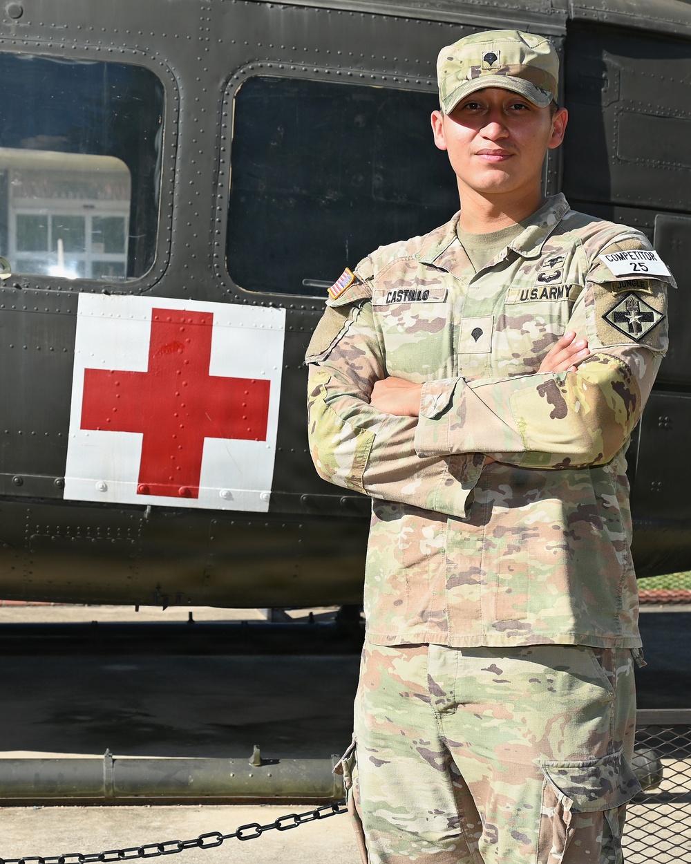 A U.S. Army soldier in OCP (Operational Camouflage Pattern) uniform stands with arms crossed in front of a military medical vehicle marked with a Red Cross symbol. The soldier's name tape reads 'CASTILLO' and wears a 'COMPETITOR 25' tag on the right sleeve, suggesting participation in a military competition. The background features what appears to be a military ambulance or medical helicopter.