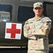 A U.S. Army soldier in OCP (Operational Camouflage Pattern) uniform stands with arms crossed in front of a military medical vehicle marked with a Red Cross symbol. The soldier's name tape reads 'CASTILLO' and wears a 'COMPETITOR 25' tag on the right sleeve, suggesting participation in a military competition. The background features what appears to be a military ambulance or medical helicopter.