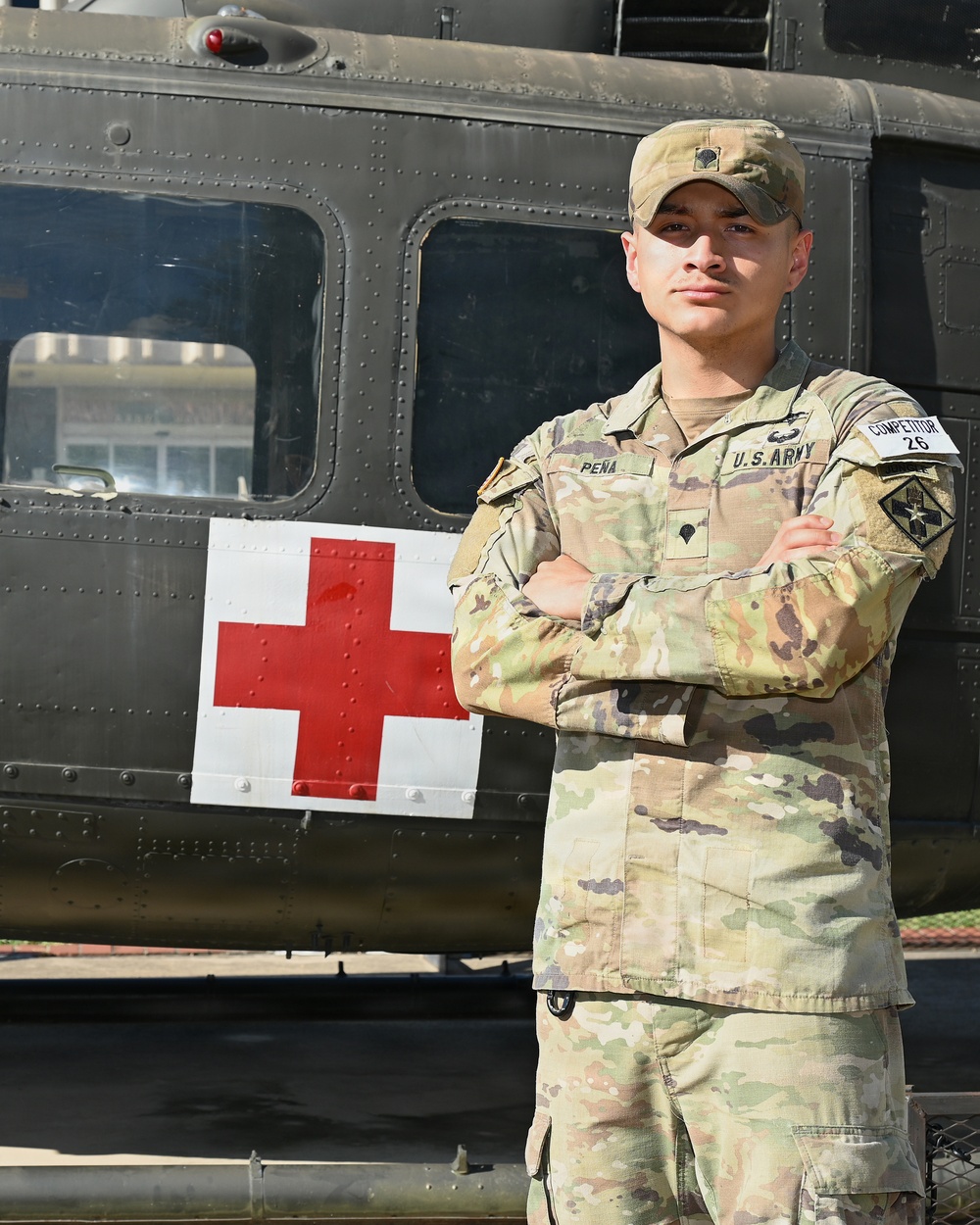 A U.S. Army soldier named Peña, wearing Operational Camouflage Pattern (OCP) uniform, stands with arms crossed in front of a military medical helicopter marked with a Red Cross symbol. The soldier is wearing a competitor bib numbered '26', suggesting participation in a military competition. The image appears to be an official or semi-official portrait-style photograph.