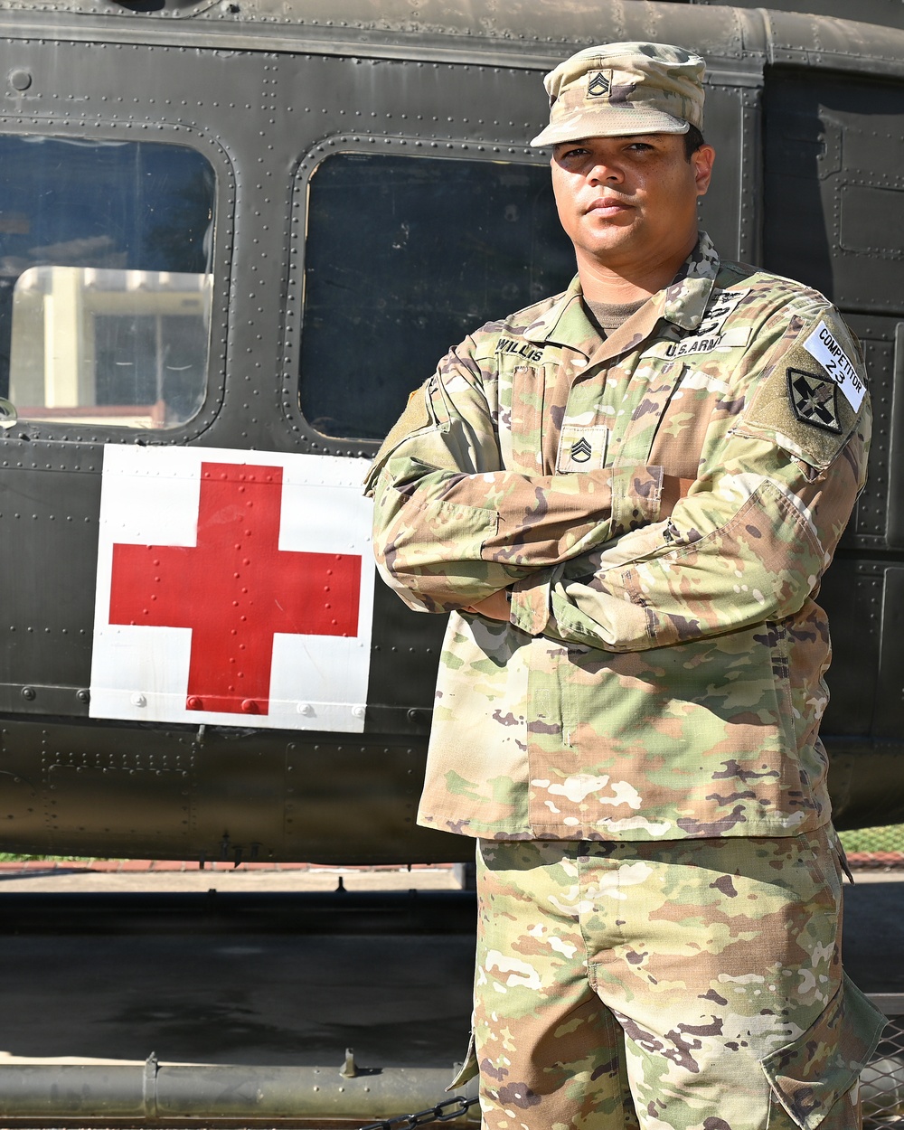 A U.S. Army Staff Sergeant named Willis stands with arms crossed in front of a military medical helicopter marked with a Red Cross symbol. He is wearing OCP (Operational Camouflage Pattern) uniform with visible rank insignia, name tape, U.S. Army tape, and a unit patch on his left shoulder. The image appears to be an official or promotional military portrait.