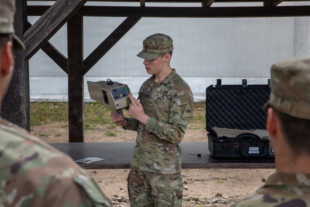 A U.S. Army soldier in OCP (Operational Camouflage Pattern) uniform is conducting a briefing or demonstration while holding a tan/beige military electronic device. The soldier is standing under a covered outdoor structure, addressing other uniformed personnel visible in the foreground. Military equipment cases are visible on a table in the background.