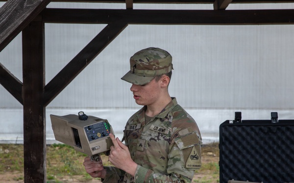 A U.S. Army soldier in OCP (Operational Camouflage Pattern) uniform is conducting a briefing or demonstration while holding a tan/beige military electronic device. The soldier is standing under a covered outdoor structure, addressing other uniformed personnel visible in the foreground. Military equipment cases are visible on a table in the background.