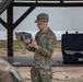 A U.S. Army soldier in OCP (Operational Camouflage Pattern) uniform is conducting a briefing or demonstration while holding a tan/beige military electronic device. The soldier is standing under a covered outdoor structure, addressing other uniformed personnel visible in the foreground. Military equipment cases are visible on a table in the background.