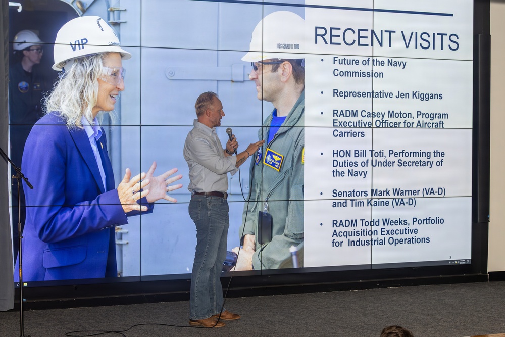 A presenter stands in front of a large display screen showing a slide titled 'Recent Visits' with bullet points listing notable Navy-related visitors. The screen displays a photograph of a woman wearing a 'VIP' hard hat labeled 'USS Gerald R. Ford' interacting with a military officer in a flight suit. The presentation appears to be a briefing related to the USS Gerald R. Ford aircraft carrier program.