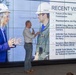 A presenter stands in front of a large display screen showing a slide titled 'Recent Visits' with bullet points listing notable Navy-related visitors. The screen displays a photograph of a woman wearing a 'VIP' hard hat labeled 'USS Gerald R. Ford' interacting with a military officer in a flight suit. The presentation appears to be a briefing related to the USS Gerald R. Ford aircraft carrier program.