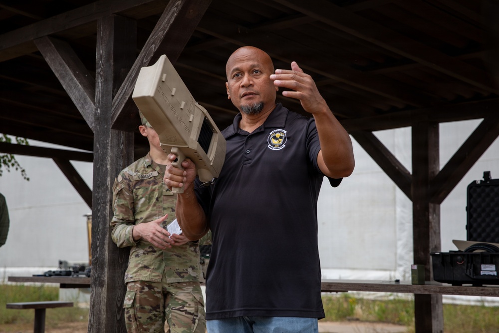 A civilian instructor or contractor wearing a black polo shirt with an emblem is holding a tan/beige military-style electronic device (likely a counter-drone or RF jamming system) while gesturing and speaking. Behind him stands a uniformed military personnel in MultiCam/OCP camouflage. The setting appears to be an outdoor covered pavilion or shelter, suggesting a training or demonstration event.