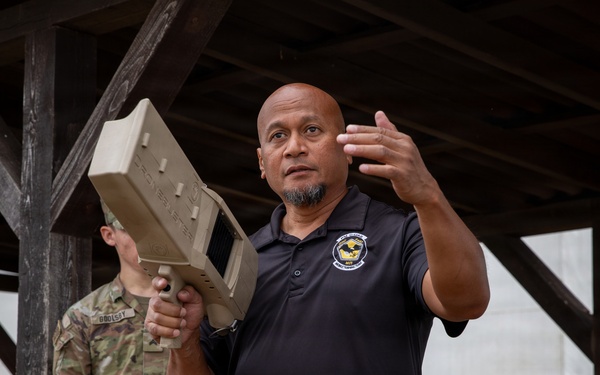 A civilian instructor or contractor wearing a black polo shirt with an emblem is holding a tan/beige military-style electronic device (likely a counter-drone or RF jamming system) while gesturing and speaking. Behind him stands a uniformed military personnel in MultiCam/OCP camouflage. The setting appears to be an outdoor covered pavilion or shelter, suggesting a training or demonstration event.