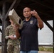 A civilian instructor or contractor wearing a black polo shirt with an emblem is holding a tan/beige military-style electronic device (likely a counter-drone or RF jamming system) while gesturing and speaking. Behind him stands a uniformed military personnel in MultiCam/OCP camouflage. The setting appears to be an outdoor covered pavilion or shelter, suggesting a training or demonstration event.
