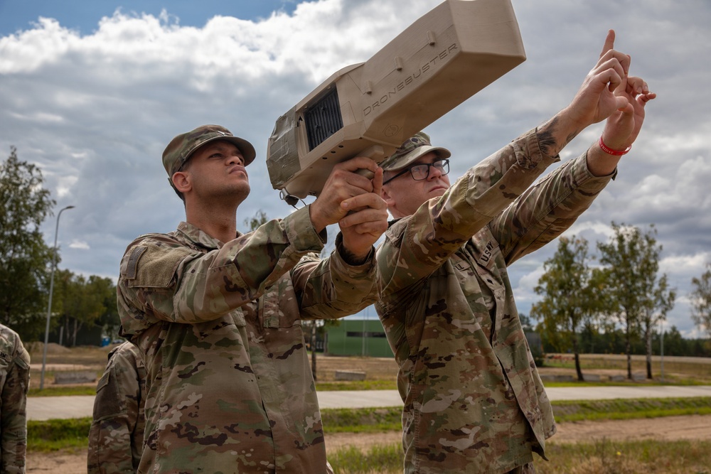 Two U.S. Army soldiers in OCP (Operational Camouflage Pattern) uniforms are handling a 'Dronebuster' counter-drone device outdoors at what appears to be a military installation. One soldier is aiming the device skyward while the other points upward, suggesting training or demonstration of the anti-drone system. The setting appears to be a European military base based on the landscape and infrastructure visible in the background.