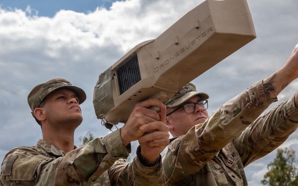 Two U.S. Army soldiers in OCP (Operational Camouflage Pattern) uniforms are handling a 'Dronebuster' counter-drone device outdoors at what appears to be a military installation. One soldier is aiming the device skyward while the other points upward, suggesting training or demonstration of the anti-drone system. The setting appears to be a European military base based on the landscape and infrastructure visible in the background.