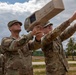 Two U.S. Army soldiers in OCP (Operational Camouflage Pattern) uniforms are handling a 'Dronebuster' counter-drone device outdoors at what appears to be a military installation. One soldier is aiming the device skyward while the other points upward, suggesting training or demonstration of the anti-drone system. The setting appears to be a European military base based on the landscape and infrastructure visible in the background.
