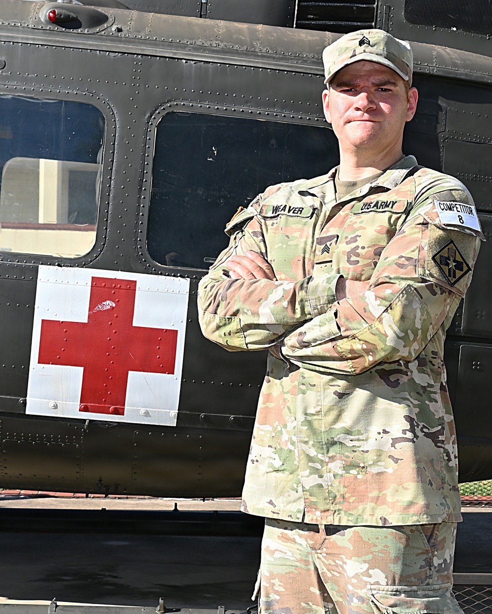 A U.S. Army Sergeant named Weaver stands with arms crossed in front of a military medical helicopter (medevac), identifiable by the prominent Red Cross marking on its fuselage. He is wearing OCP (Operational Camouflage Pattern) uniform with a 'COMPETITOR 8' armband on his right sleeve, suggesting participation in a military competition. The image appears to be an official or semi-official portrait-style photograph.