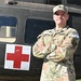 A U.S. Army Sergeant named Weaver stands with arms crossed in front of a military medical helicopter (medevac), identifiable by the prominent Red Cross marking on its fuselage. He is wearing OCP (Operational Camouflage Pattern) uniform with a 'COMPETITOR 8' armband on his right sleeve, suggesting participation in a military competition. The image appears to be an official or semi-official portrait-style photograph.