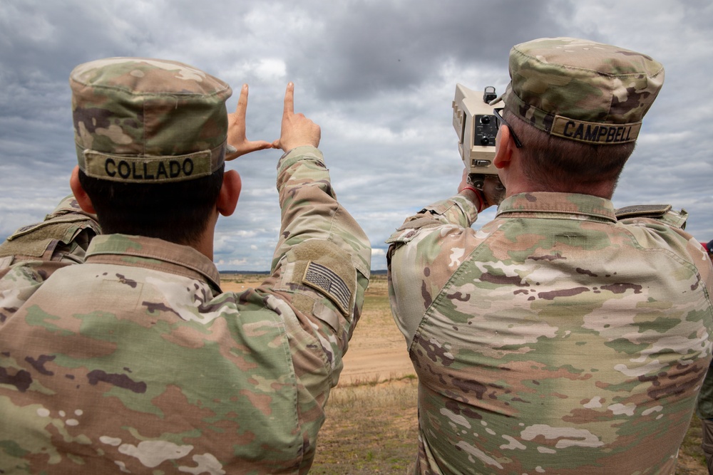 Two U.S. Army soldiers in Operational Camouflage Pattern (OCP) uniforms are conducting field operations, with one soldier operating what appears to be a laser rangefinder or targeting device while the other uses hand signals to indicate direction or distance. Their name tapes read 'COLLADO' and 'CAMPBELL'. The setting appears to be an open training range or field environment under overcast skies.