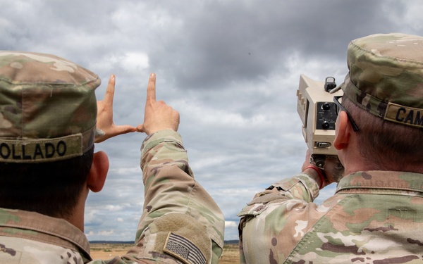 Two U.S. Army soldiers in Operational Camouflage Pattern (OCP) uniforms are conducting field operations, with one soldier operating what appears to be a laser rangefinder or targeting device while the other uses hand signals to indicate direction or distance. Their name tapes read 'COLLADO' and 'CAMPBELL'. The setting appears to be an open training range or field environment under overcast skies.