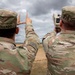 Two U.S. Army soldiers in Operational Camouflage Pattern (OCP) uniforms are conducting field operations, with one soldier operating what appears to be a laser rangefinder or targeting device while the other uses hand signals to indicate direction or distance. Their name tapes read 'COLLADO' and 'CAMPBELL'. The setting appears to be an open training range or field environment under overcast skies.