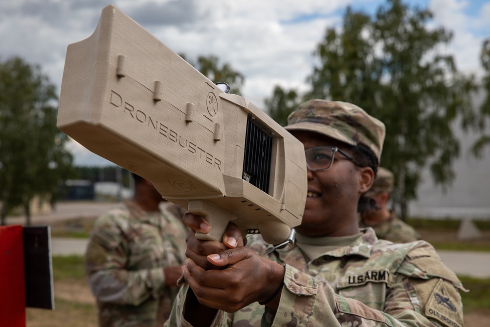 A U.S. Army soldier in OCP (Operational Camouflage Pattern) uniform is holding and aiming a 'Dronebuster' counter-drone electronic warfare device. The soldier is smiling and wearing protective eyewear, suggesting a training or demonstration context. Additional soldiers in uniform are visible in the background at what appears to be an outdoor military installation.