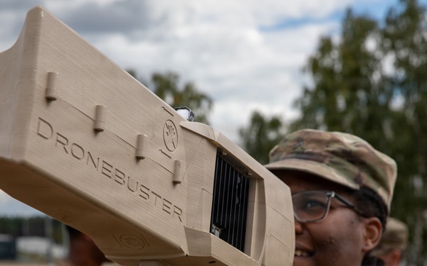 A U.S. Army soldier in OCP (Operational Camouflage Pattern) uniform is holding and aiming a 'Dronebuster' counter-drone electronic warfare device. The soldier is smiling and wearing protective eyewear, suggesting a training or demonstration context. Additional soldiers in uniform are visible in the background at what appears to be an outdoor military installation.