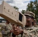 A U.S. Army soldier in OCP (Operational Camouflage Pattern) uniform is holding and aiming a 'Dronebuster' counter-drone electronic warfare device. The soldier is smiling and wearing protective eyewear, suggesting a training or demonstration context. Additional soldiers in uniform are visible in the background at what appears to be an outdoor military installation.
