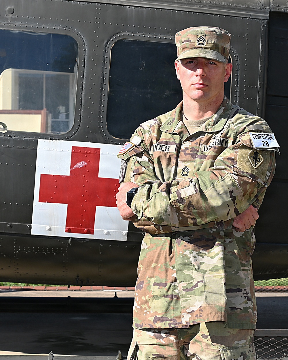 A U.S. Army Staff Sergeant in MultiCam/OCP camouflage uniform stands with arms crossed in front of a military medical helicopter marked with a Red Cross symbol. The soldier wears a 'COMPETITOR 28' tab on the right sleeve, suggesting participation in a military competition. The image appears to be an official or semi-official portrait-style photograph.