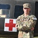 A U.S. Army Staff Sergeant in MultiCam/OCP camouflage uniform stands with arms crossed in front of a military medical helicopter marked with a Red Cross symbol. The soldier wears a 'COMPETITOR 28' tab on the right sleeve, suggesting participation in a military competition. The image appears to be an official or semi-official portrait-style photograph.