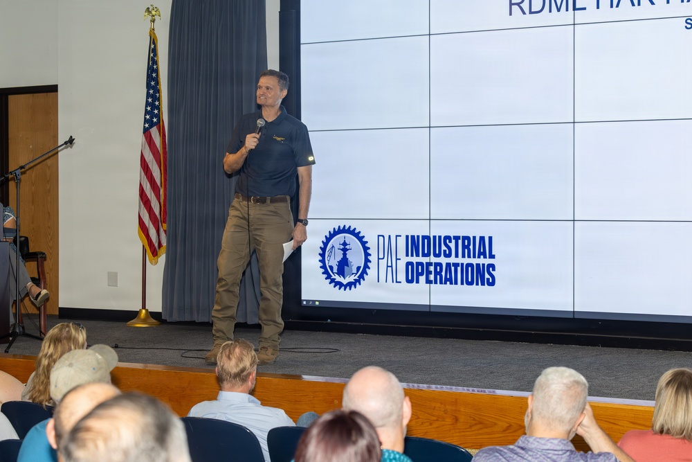 A man is delivering a presentation or speech at a formal gathering, standing in front of a large projection screen displaying the 'PAE Industrial Operations' logo. An American flag is positioned to his left, and an audience is seated facing him. The setting appears to be a conference room or auditorium within a military or defense-related facility.