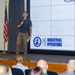 A man is delivering a presentation or speech at a formal gathering, standing in front of a large projection screen displaying the 'PAE Industrial Operations' logo. An American flag is positioned to his left, and an audience is seated facing him. The setting appears to be a conference room or auditorium within a military or defense-related facility.