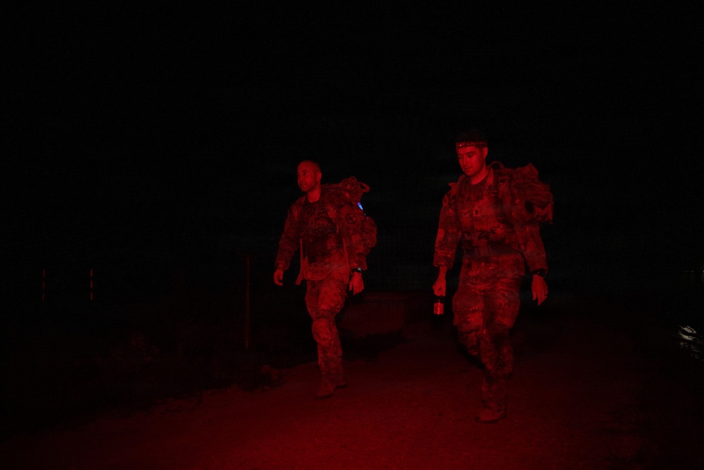 Two military personnel in camouflage uniforms are walking at night, illuminated by red light. They are carrying full tactical backpacks and one individual is holding a red-light flashlight or signal device. The scene appears to be a nighttime patrol or ruck march exercise.