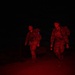 Two military personnel in camouflage uniforms are walking at night, illuminated by red light. They are carrying full tactical backpacks and one individual is holding a red-light flashlight or signal device. The scene appears to be a nighttime patrol or ruck march exercise.