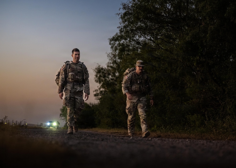 Two military personnel in camouflage uniforms and tactical gear are walking along a gravel path at dusk or dawn. They are moving toward the camera with vegetation on the right side and vehicle headlights visible in the background. The low-light conditions create a dramatic silhouette-like atmosphere.