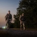 Two military personnel in camouflage uniforms and tactical gear are walking along a gravel path at dusk or dawn. They are moving toward the camera with vegetation on the right side and vehicle headlights visible in the background. The low-light conditions create a dramatic silhouette-like atmosphere.
