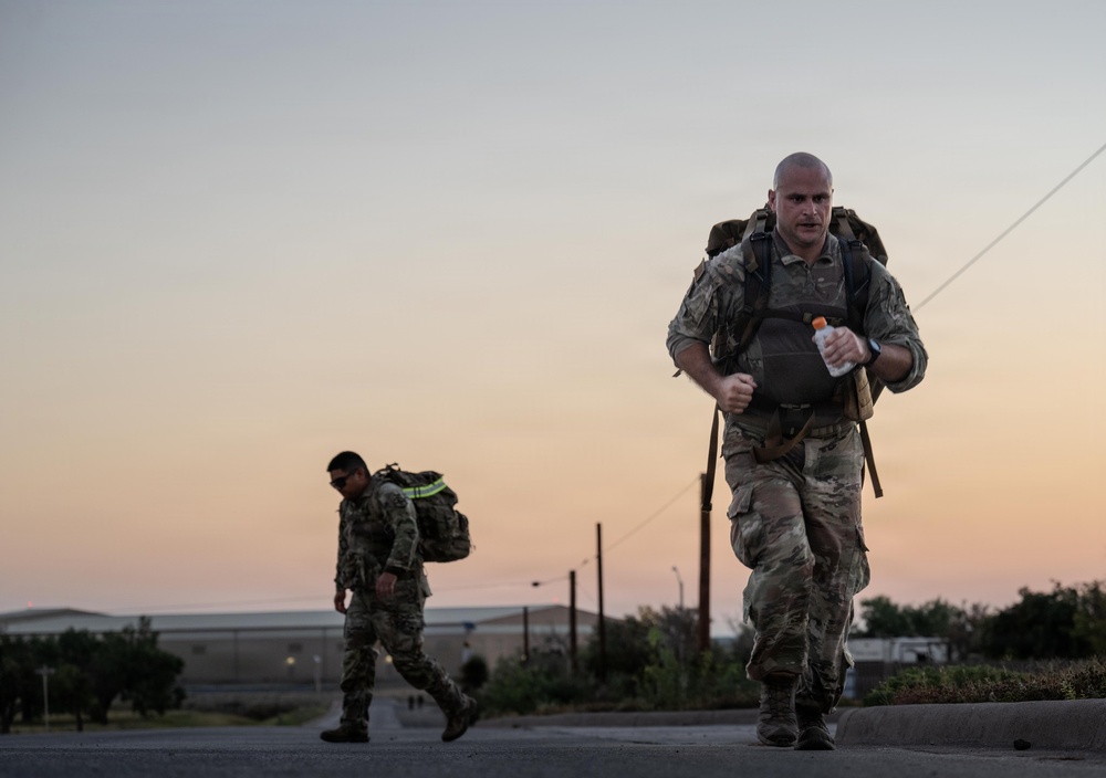 Two military personnel in camouflage uniforms are conducting a ruck march or physical training exercise at dawn or dusk. The foreground soldier is running while carrying a loaded backpack and holding a water bottle, while a second soldier walks behind. The setting appears to be a military installation road with a colorful twilight sky in the background.