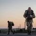 Two military personnel in camouflage uniforms are conducting a ruck march or physical training exercise at dawn or dusk. The foreground soldier is running while carrying a loaded backpack and holding a water bottle, while a second soldier walks behind. The setting appears to be a military installation road with a colorful twilight sky in the background.