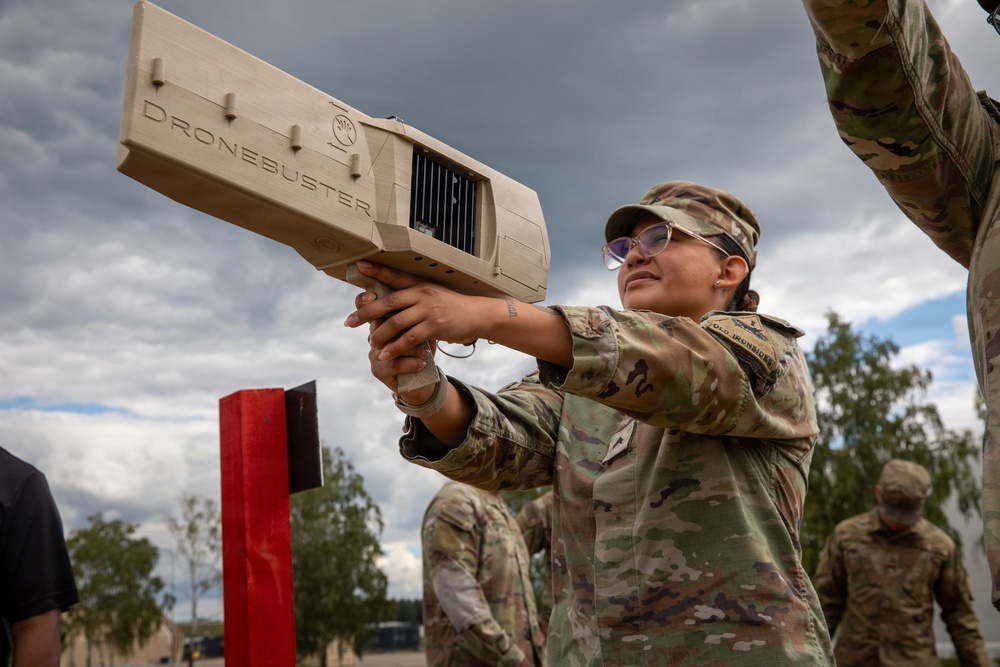A female soldier in MultiCam/OCP camouflage uniform is aiming a 'Dronebuster' counter-drone electronic warfare device skyward during what appears to be a training exercise. The Dronebuster is a handheld radio frequency (RF) jamming system designed to neutralize unmanned aerial systems (UAS). Additional soldiers in matching uniforms are visible in the background at what appears to be a military training area.