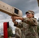 A female soldier in MultiCam/OCP camouflage uniform is aiming a 'Dronebuster' counter-drone electronic warfare device skyward during what appears to be a training exercise. The Dronebuster is a handheld radio frequency (RF) jamming system designed to neutralize unmanned aerial systems (UAS). Additional soldiers in matching uniforms are visible in the background at what appears to be a military training area.