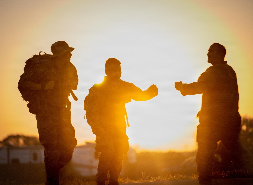 Three individuals in military camouflage uniforms are silhouetted against a vivid golden sunset. Two of the figures appear to be performing a fist bump or elbow bump greeting gesture. The warm backlit composition creates a dramatic silhouette effect with strong orange and yellow tones dominating the background.