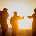 Three individuals in military camouflage uniforms are silhouetted against a vivid golden sunset. Two of the figures appear to be performing a fist bump or elbow bump greeting gesture. The warm backlit composition creates a dramatic silhouette effect with strong orange and yellow tones dominating the background.