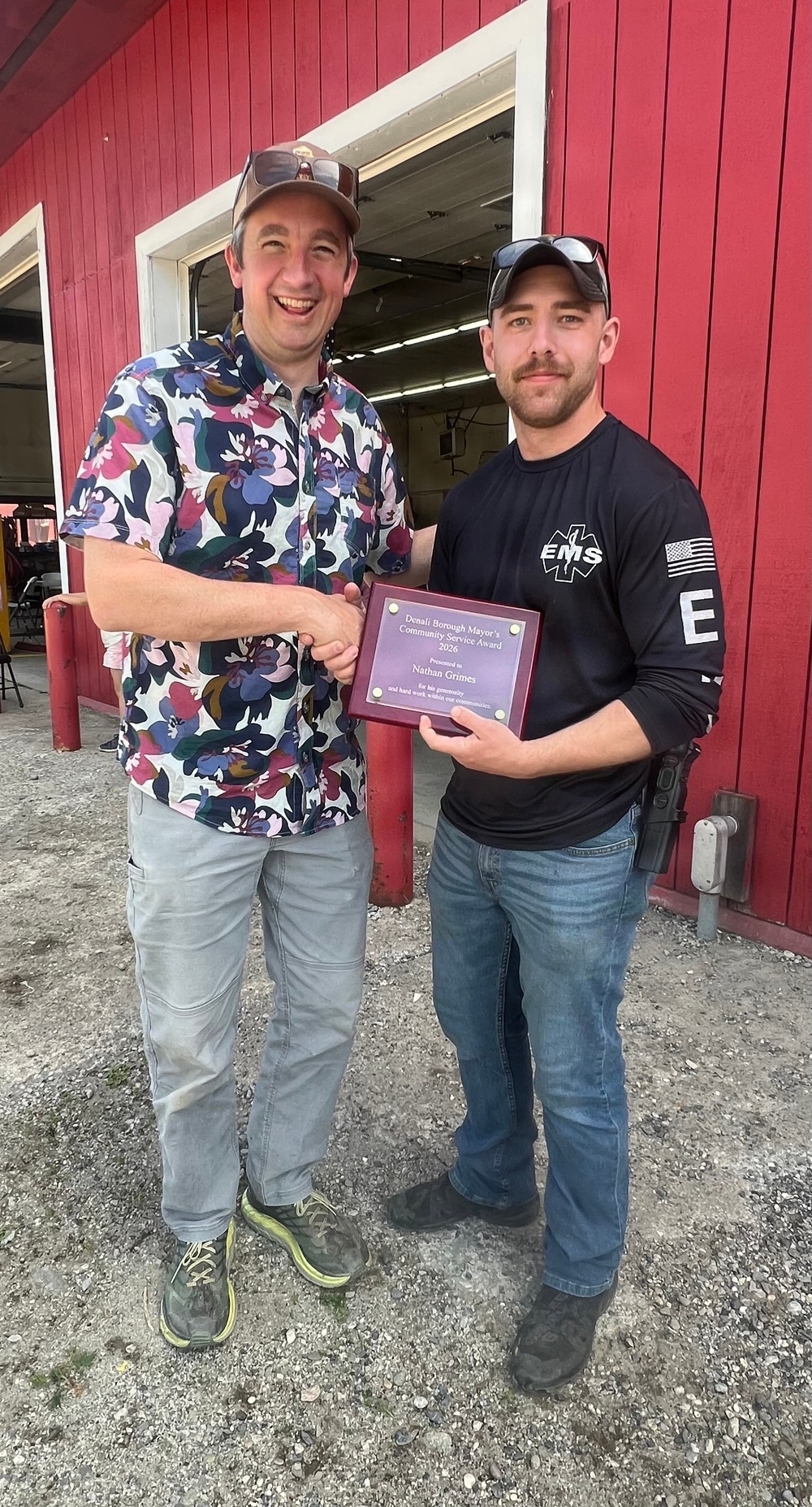 Two men are posing together outdoors in front of a red building, shaking hands while jointly holding a plaque. The plaque appears to be a 'Denali Borough Mayor's Community Service Award 2026' presented to 'Nathan Grimes.' The man on the right is wearing an EMS shirt with a US flag patch, suggesting he is an emergency medical services worker being recognized for community service.