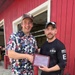 Two men are posing together outdoors in front of a red building, shaking hands while jointly holding a plaque. The plaque appears to be a 'Denali Borough Mayor's Community Service Award 2026' presented to 'Nathan Grimes.' The man on the right is wearing an EMS shirt with a US flag patch, suggesting he is an emergency medical services worker being recognized for community service.