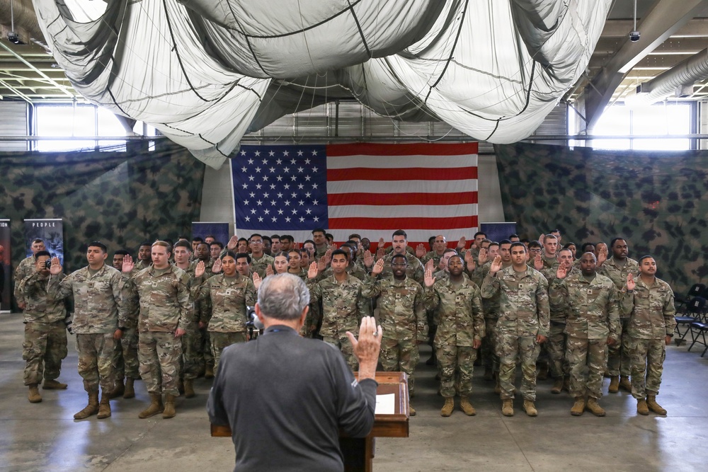 A military re-enlistment or oath-of-office ceremony is taking place inside a large hangar or military facility. A civilian official stands at a podium with his right hand raised, administering an oath to a large group of uniformed soldiers who have their right hands raised. An American flag is prominently displayed on the wall behind the assembled troops.