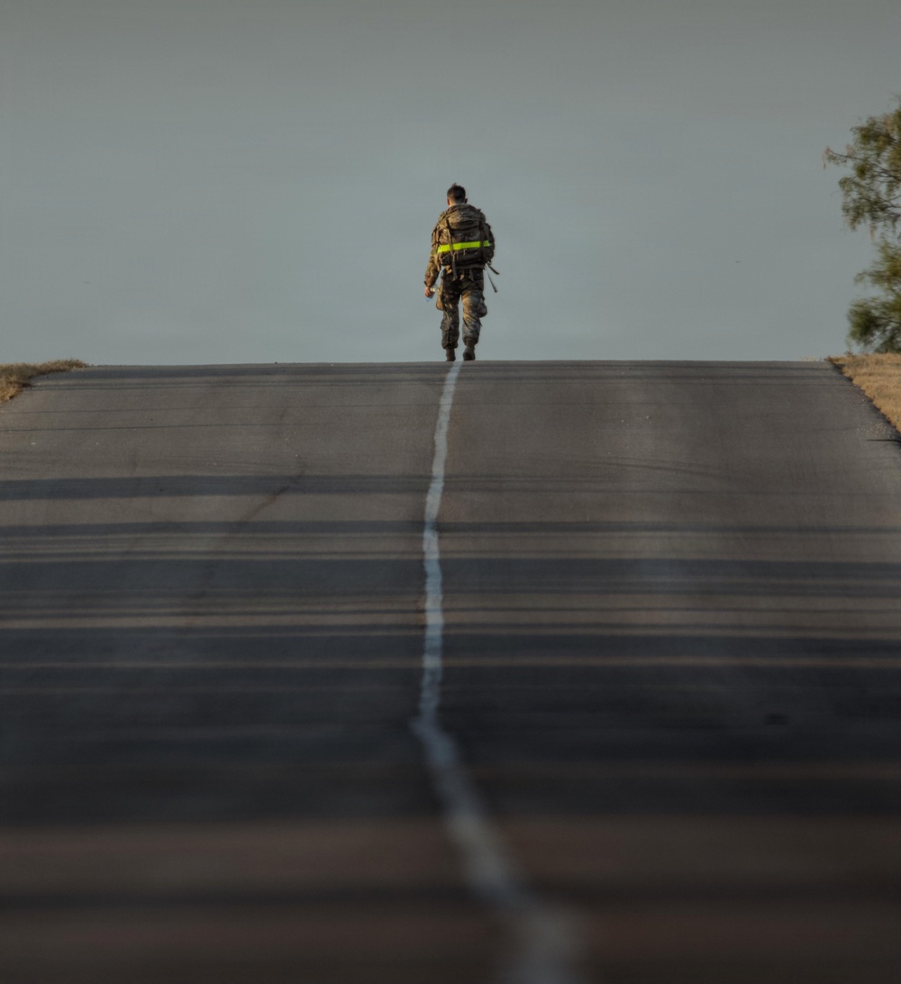 A single figure in military/tactical gear walks away from the camera along the center line of a straight, empty road. The person is carrying a large backpack with a prominent yellow-green reflective strip. The image uses shallow depth of field, with the foreground road blurred and the subject in sharp focus near the horizon.